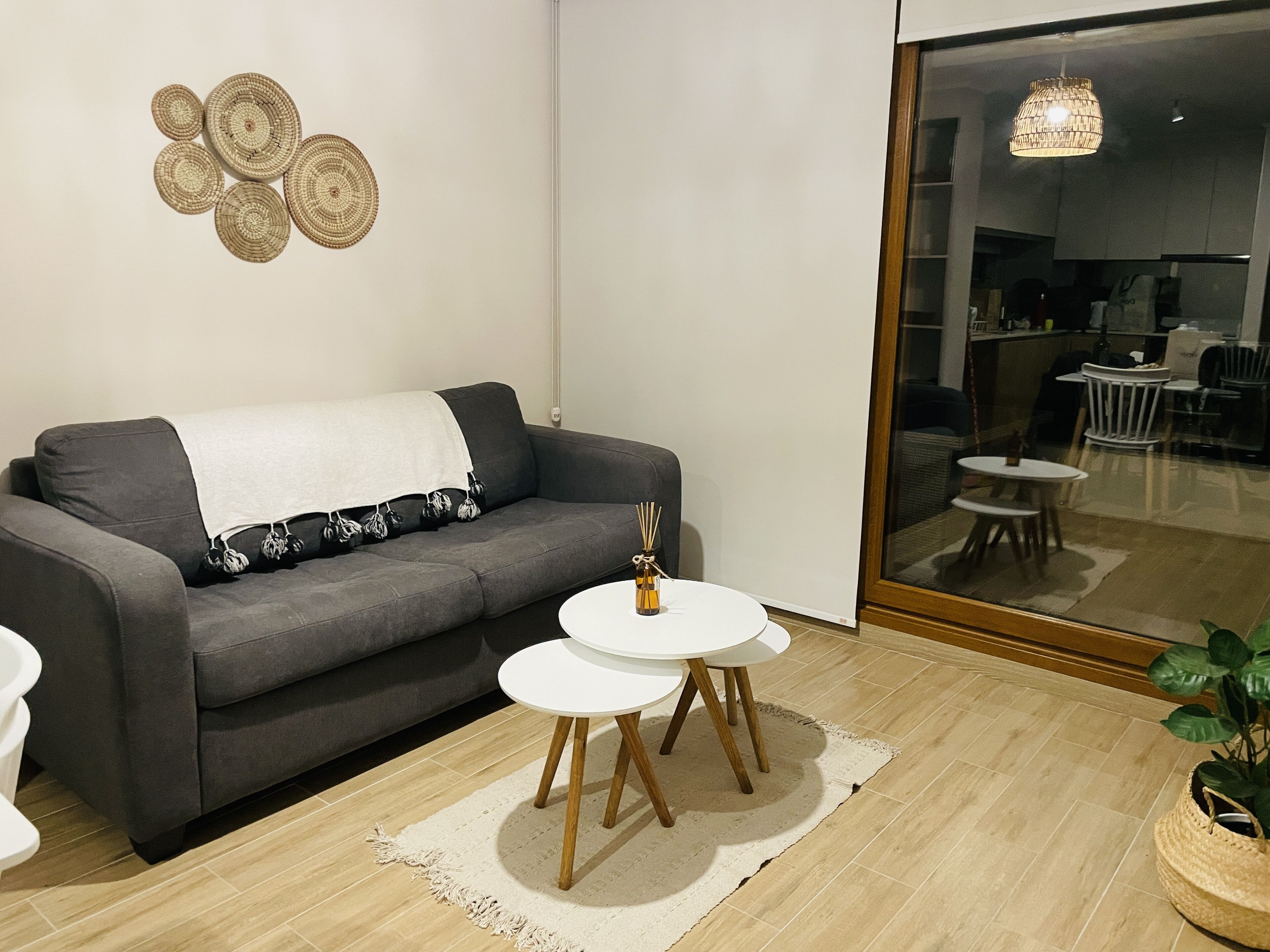 Ett modernt vardagsrum med en grå soffa, två vita, runda bord, en vävd väggdekoration och ett stort fönster med utsikt över en matplats.