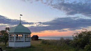En liten, envåningsbyggnad med ett grönt, kupolformat tak och en flaggstång, belägen på en gräsyta med utsikt över solnedgången över vattnet.