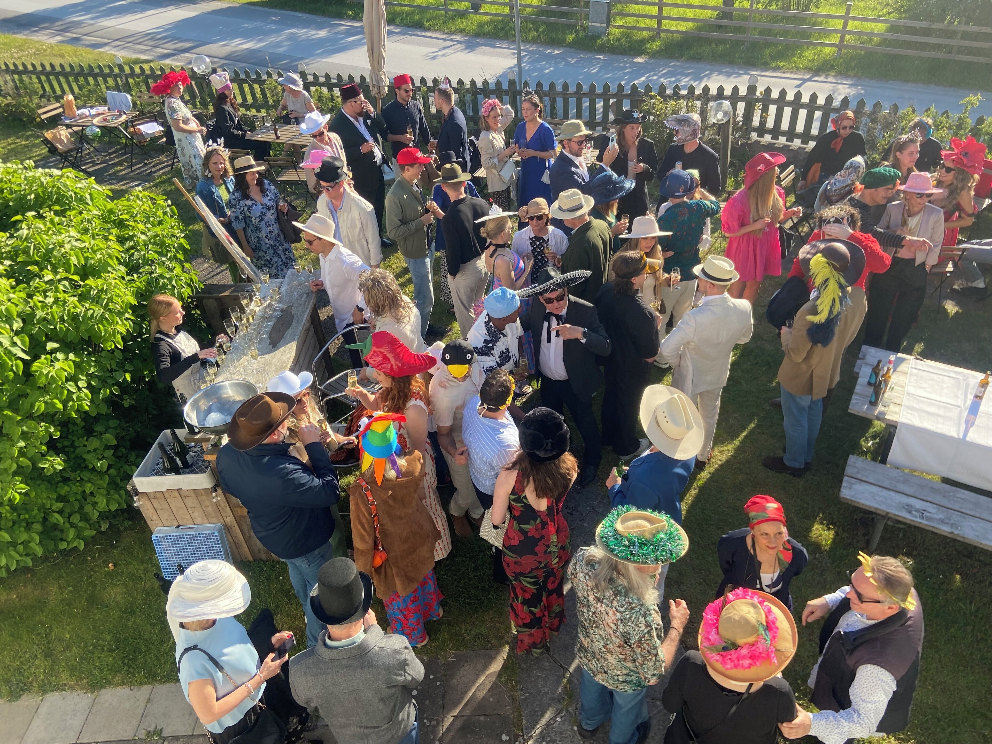 A group of people gathered outdoors, some wearing hats and casual clothing, with tables and chairs visible.
