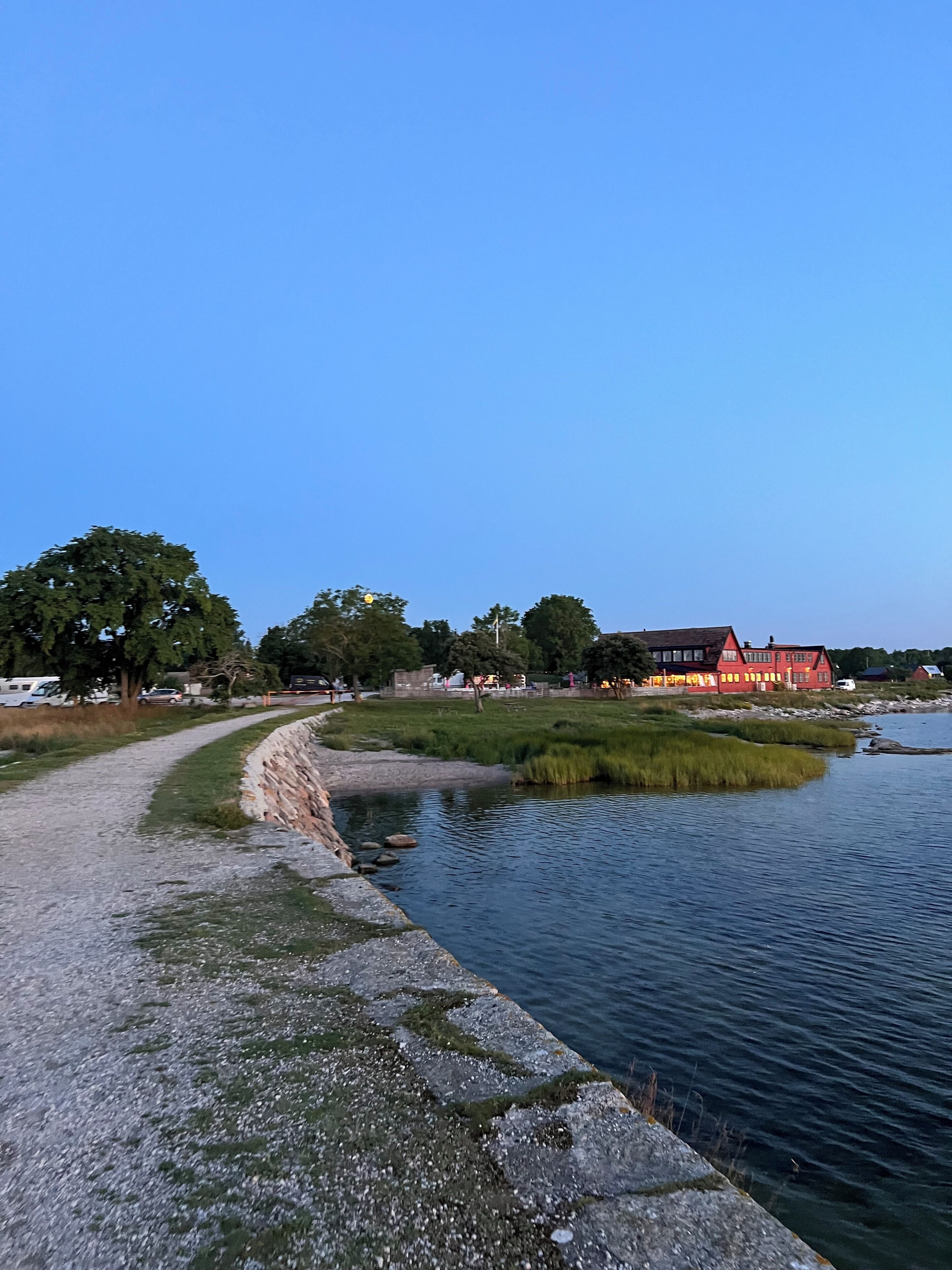 A riverside path with a stone wall, a red building, and trees in the background.