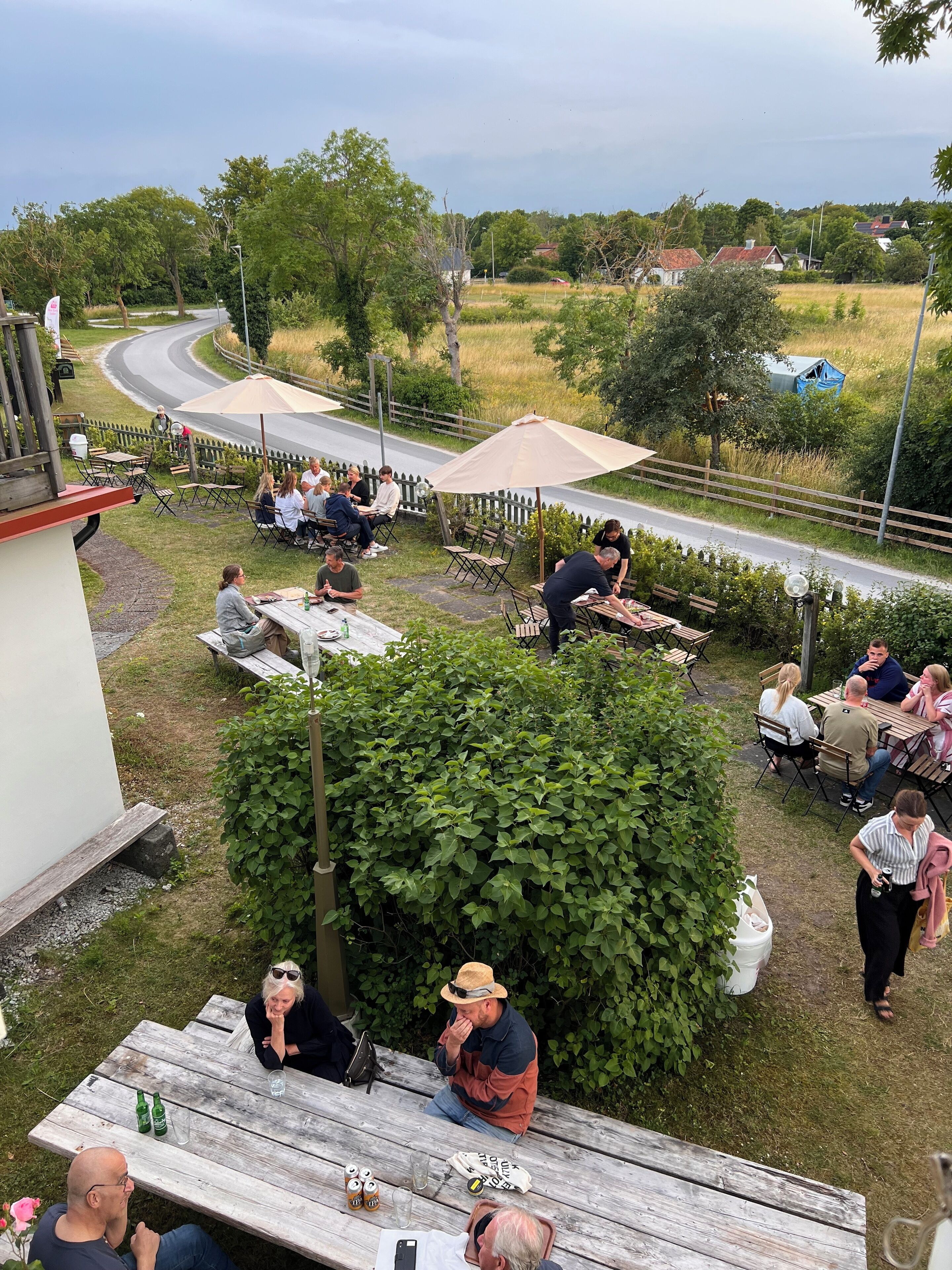 An outdoor seating area with tables and chairs, surrounded by greenery and a view of a road.
