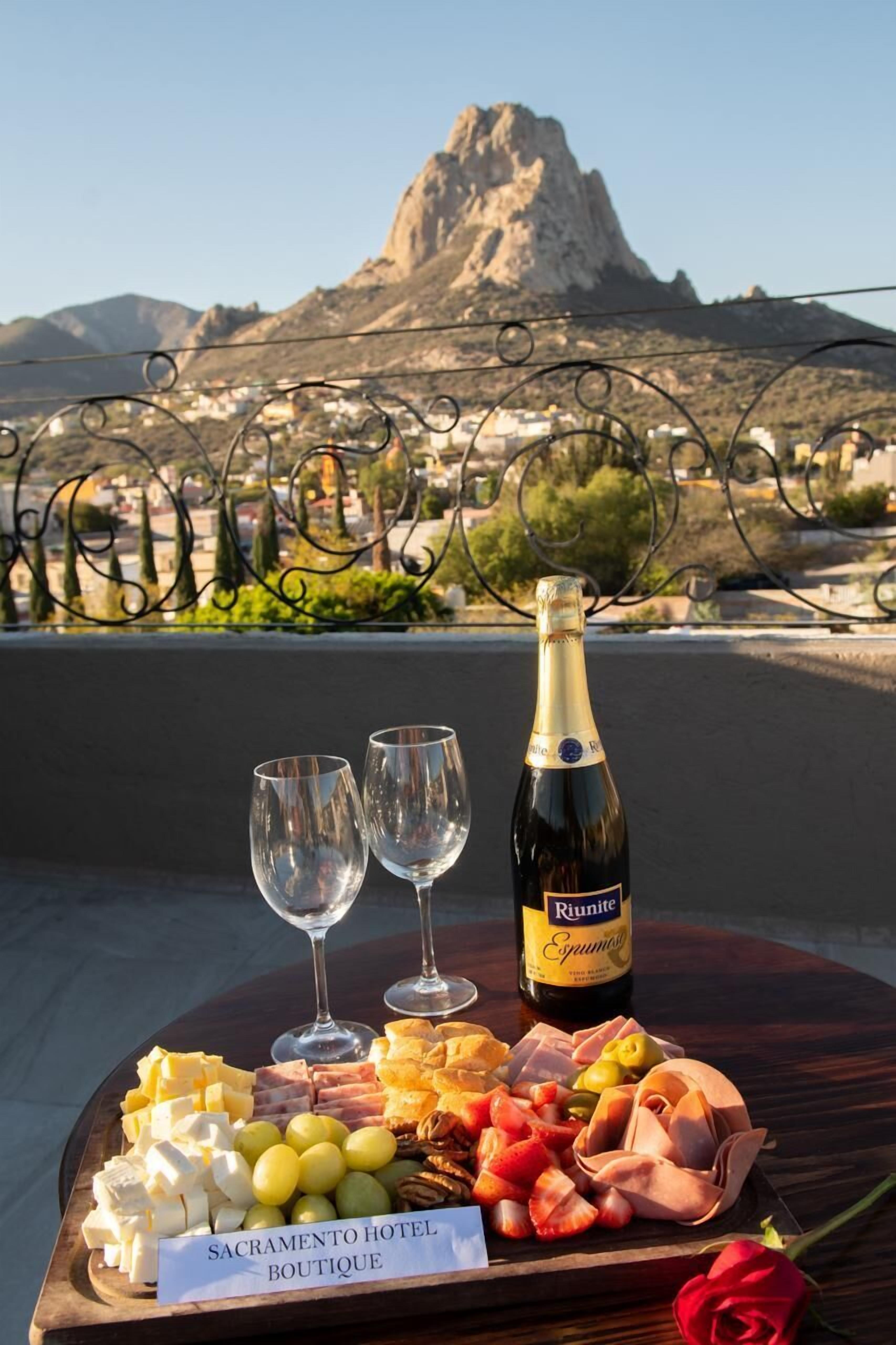 A table with a bottle of Riumite Espumante, two glasses, and a variety of cheeses, fruits, and meats. A sign reads "Sacramento Hotel Boutique."