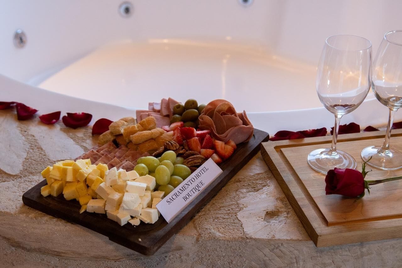 A buffet with various cheeses, fruits, and meats, accompanied by two wine glasses and a rose on a tray.
