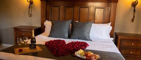 A neatly made bed with a wooden headboard, two bedside tables, and a tray with a bottle, glasses, and a heart-shaped arrangement of red flowers.