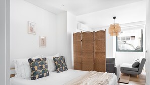 A modern bedroom with a large bed, a grey armchair, a woven partition, and a hanging light fixture. - Estrela Delight by Homing (Lisbon)
