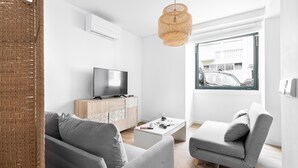 A modern living room with a grey sofa, a grey armchair, a white coffee table, a TV on a wooden cabinet, and a large window with a view of buildings. - Estrela Delight by Homing (Lisbon)