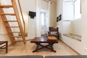 A room with a wooden table, a chair, a ladder, a remote control, and a window with a view of a door and a wall-mounted TV.