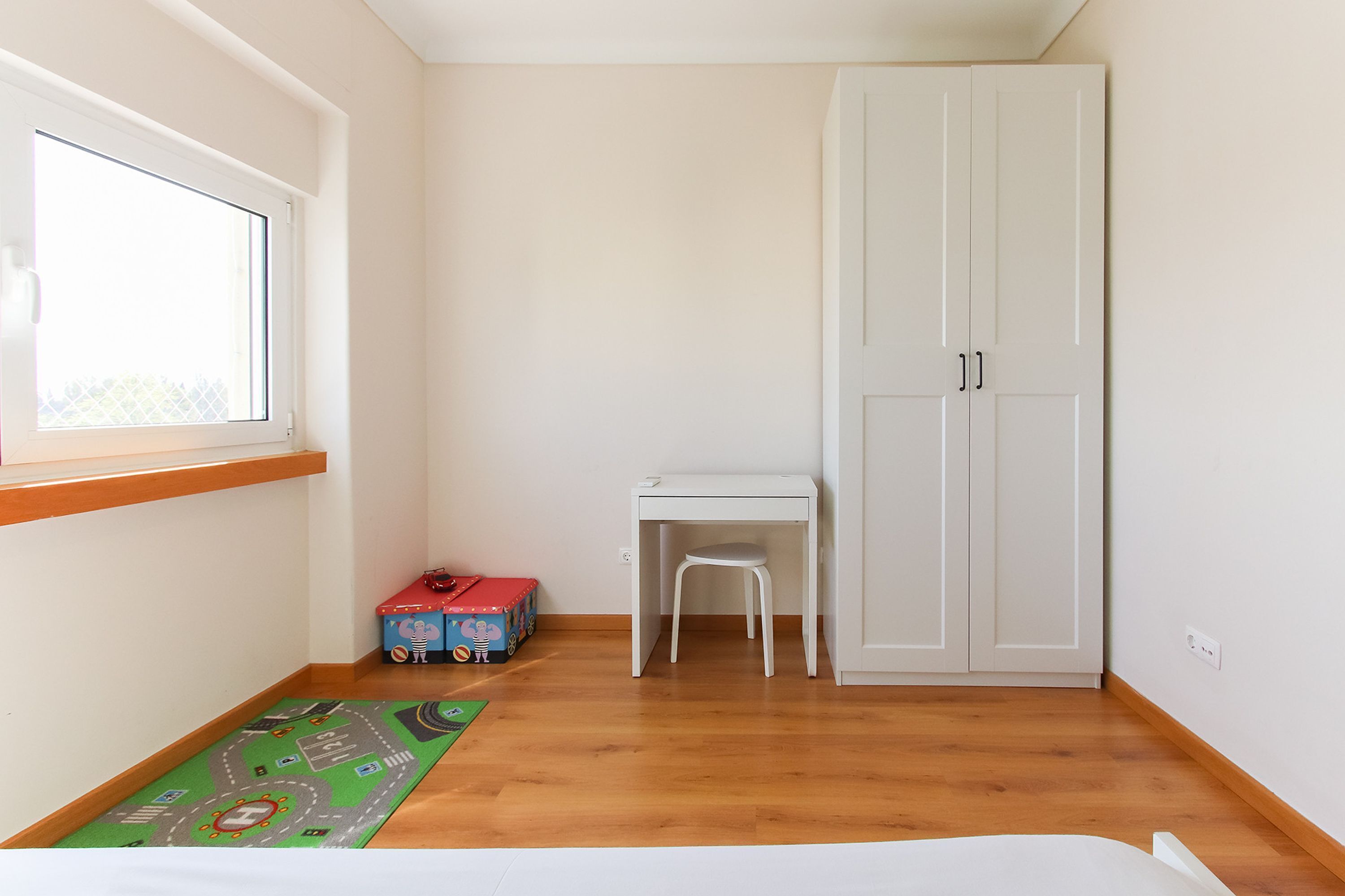 A bedroom with a wooden floor, a bed, a window, a desk, a chair, and a wardrobe.