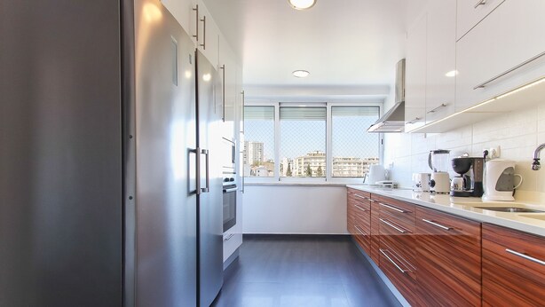 A modern kitchen with stainless steel appliances, wooden cabinets, and a large window offering a city view. - Olivais Balcony by Homing (Lisbon)