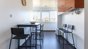A modern kitchen with a dining area, featuring a large window, wooden cabinets, and black chairs. - Olivais Balcony by Homing (Lisbon)