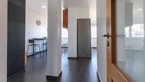 A modern interior with a dark floor, white walls, and a wooden door. There is a refrigerator, a clock on the wall, and a dining area with chairs and a table. - Olivais Balcony by Homing (Lisbon)