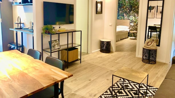 Um espaço de estar moderno com uma mesa de jantar de madeira, um suporte de TV de metal preto, uma pequena mesa redonda e um tapete estampado.
