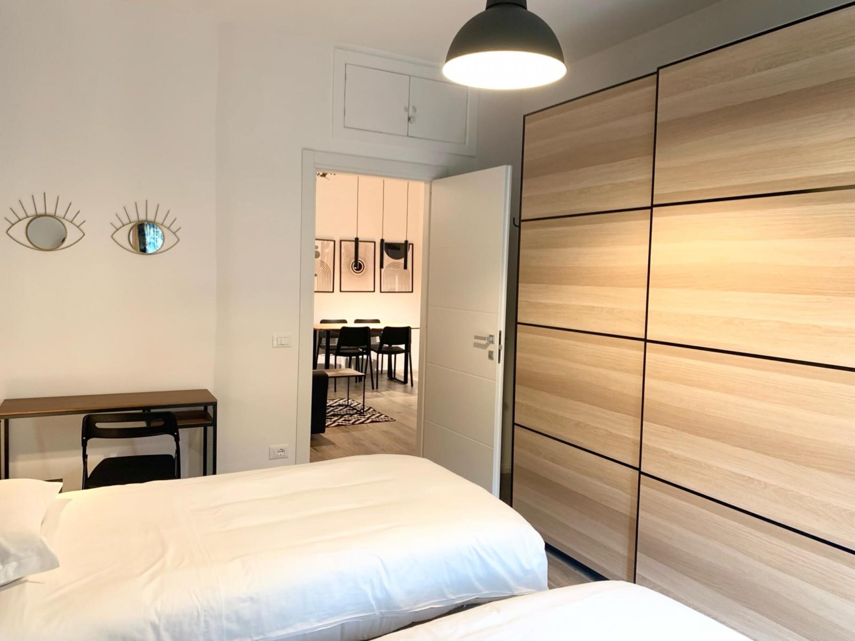 A bedroom with a bed, a desk, and a wardrobe. There is a dining area visible through an open door.