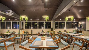 A restaurant with multiple tables and chairs, wooden ceiling, hanging plants, and large windows. - Harmony Palace (Mahabaleshwar)