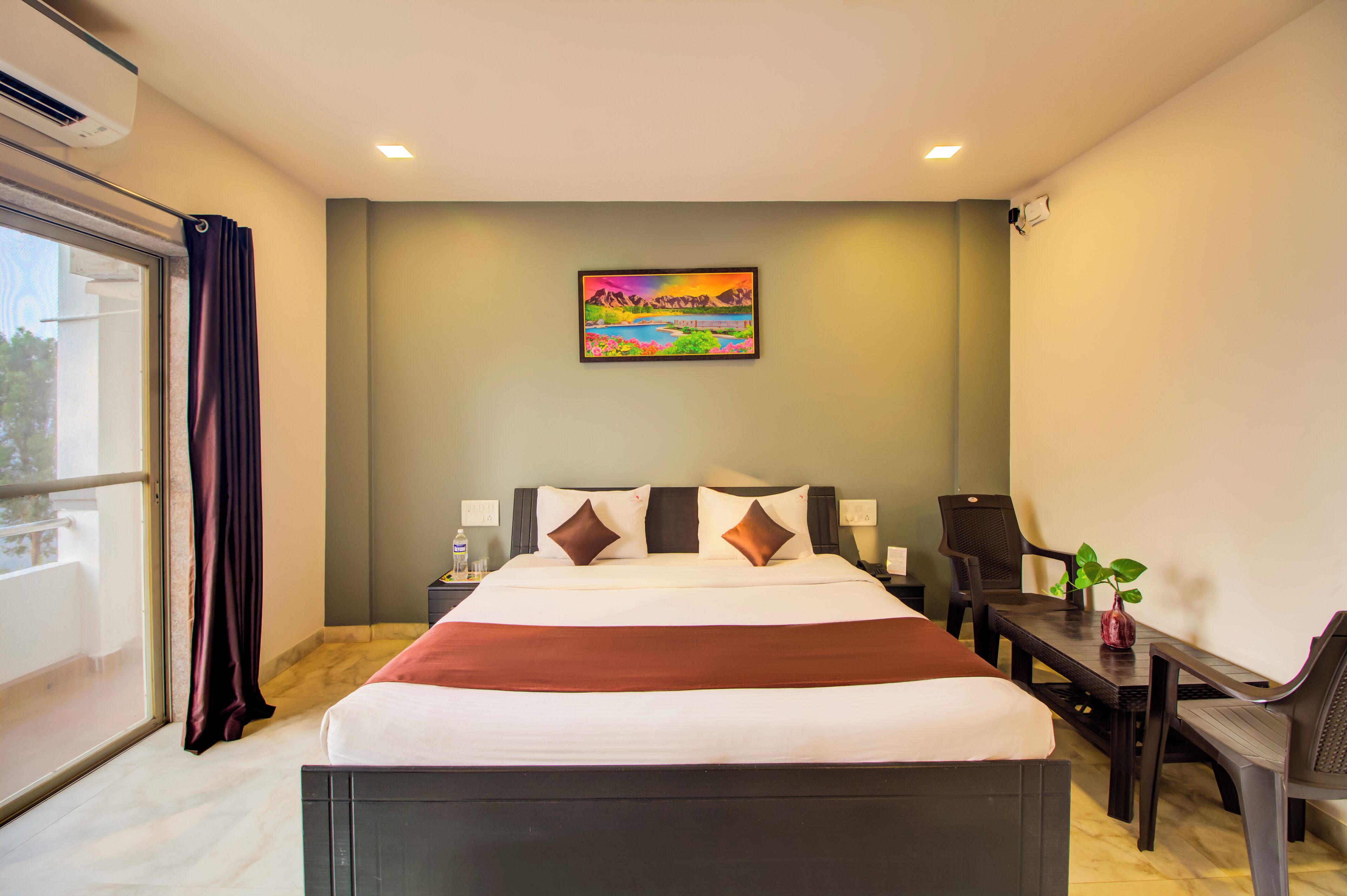 A hotel room with a bed, a desk, a chair, a potted plant, and a painting on the wall.