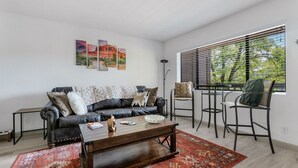 A living room with a dark leather sofa, a wooden coffee table, and a red patterned rug. There are two chairs and a floor lamp. The room has a large window with blinds, and artwork is displayed on the wall. - Sedona Eve

 (Sedona)