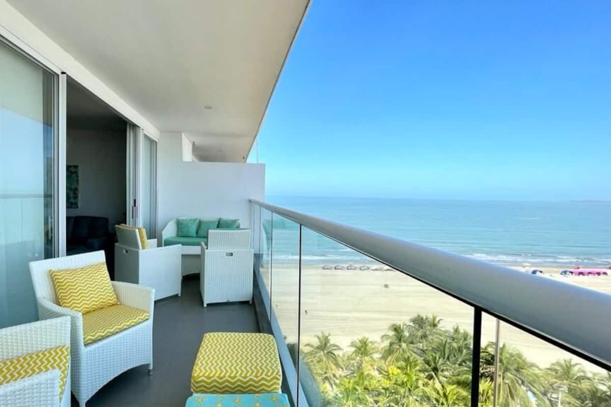 A balcony with white wicker furniture, a view of the beach, and a clear sky.