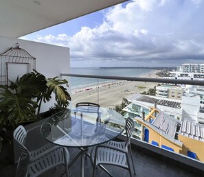 A balcony with a glass table and chairs, overlooking a beach and buildings.