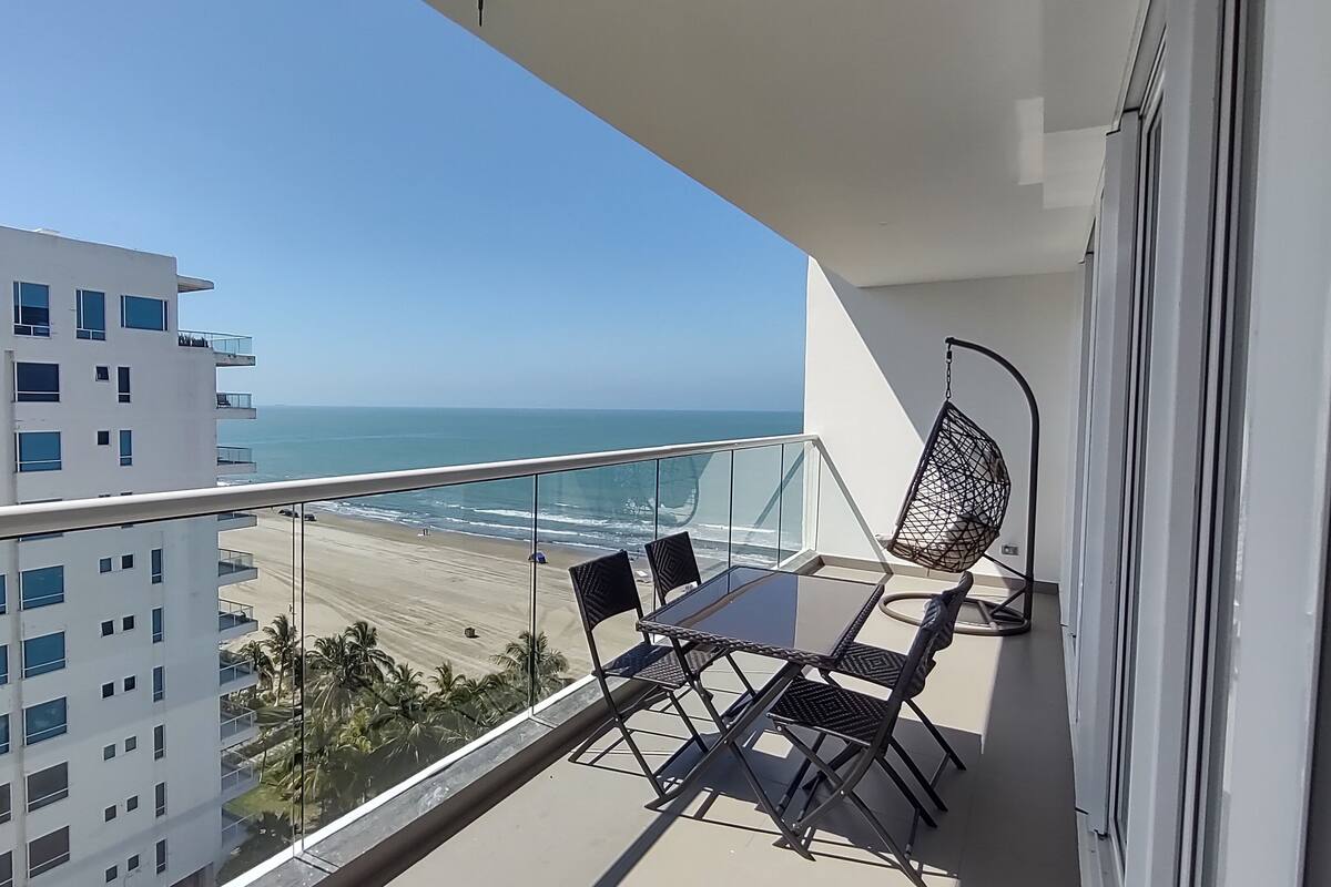 A balcony with a table and chairs, overlooking a beach and ocean.