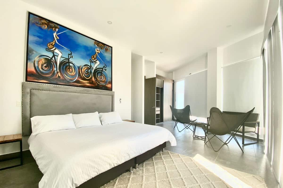 A modern hotel room with a large bed, a painting of a woman on a bicycle, a desk with chairs, and a view of the hallway.