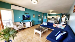 Sala de estar con sofá, mesa de centro y televisor, además de una cocina y un comedor con sillas y mesa.