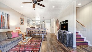 A spacious living room with a sofa, coffee table, and a red leather ottoman. A dining area with a wooden table and chairs is visible in the background. - Parkview at Red River (Red River)