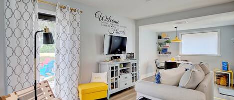 A modern living room with a sofa, a yellow ottoman, a TV, and a wall-mounted shelf with books and decorative items.