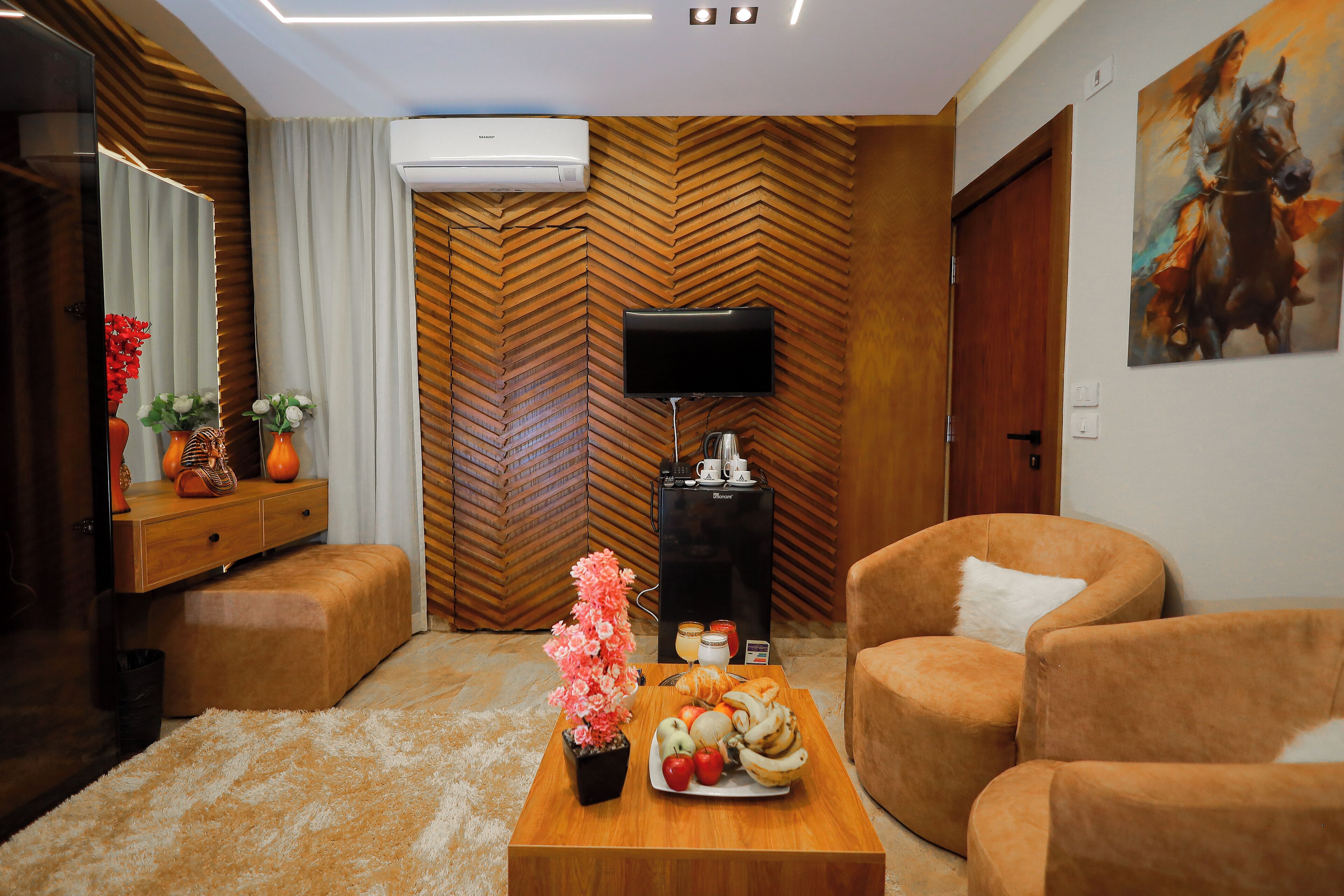 A modern living room with a wooden herringbone wall, a flat-screen TV, a coffee table with fruits and flowers, and two plush armchairs.
