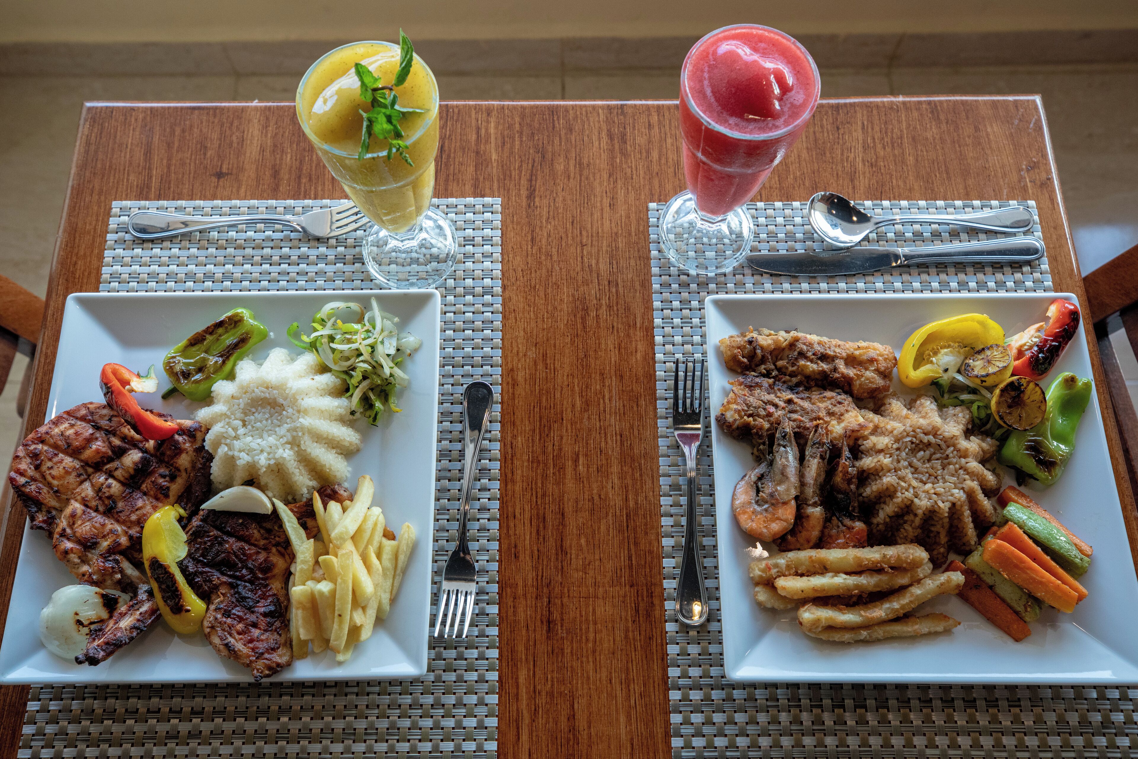 Two square plates of food with cutlery and glasses on a table.