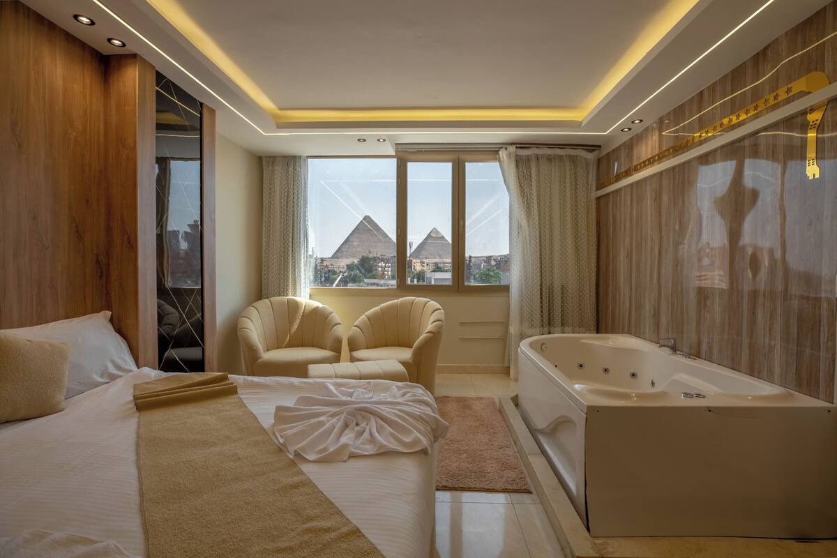 A modern hotel room with a large bed, two armchairs, a bathtub, and a view of pyramids.