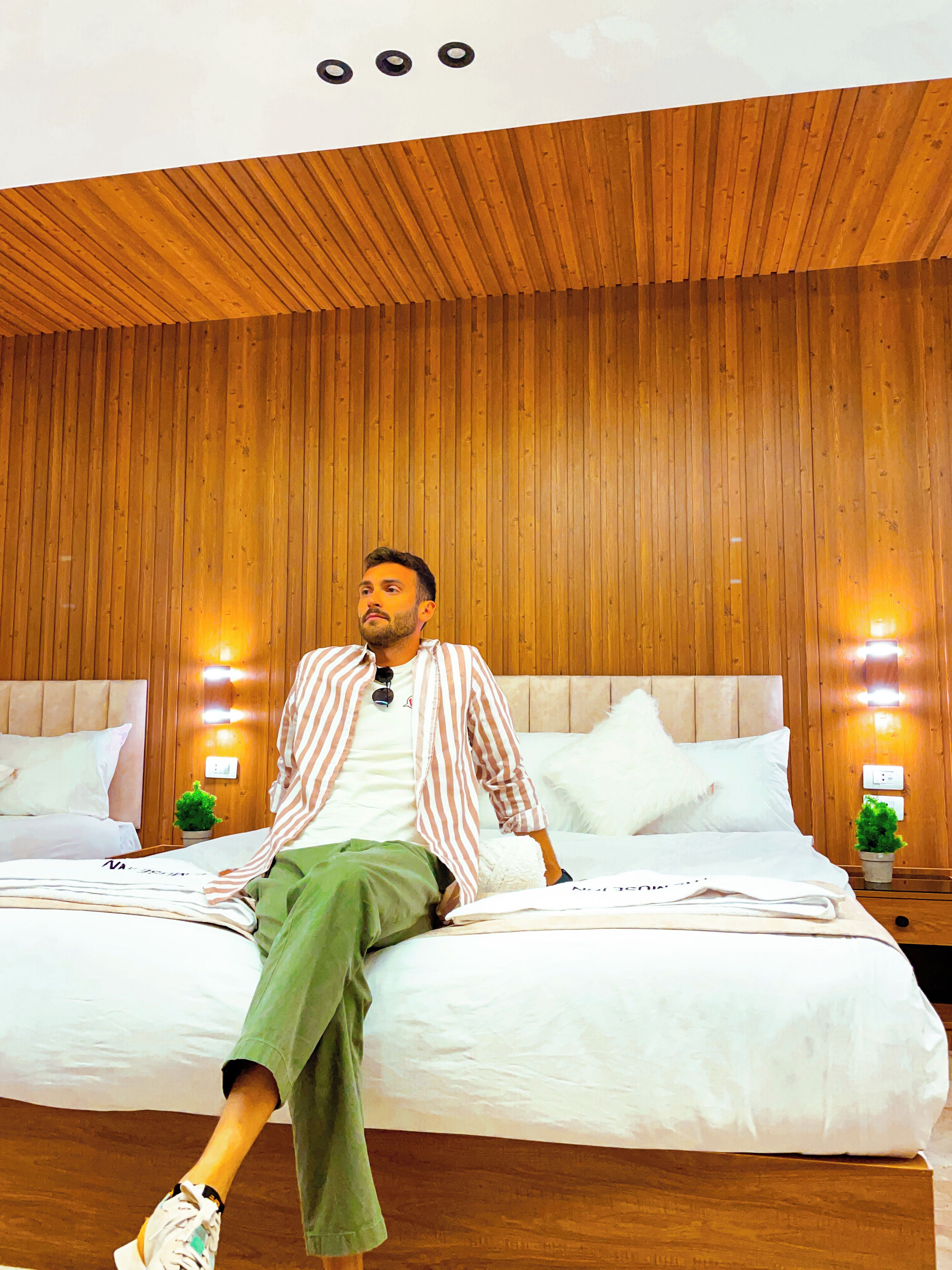 A person sitting on a bed in a room with wooden paneling and two wall-mounted lights.