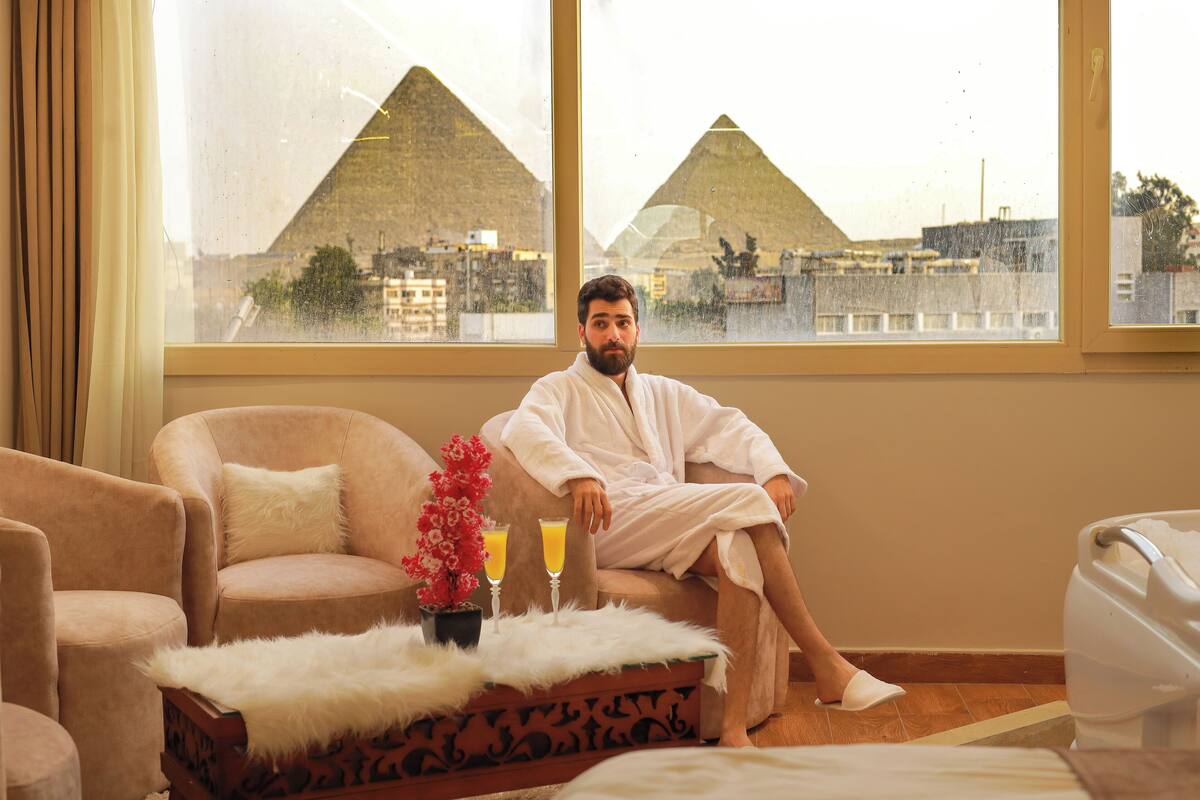 A man in a white bathrobe sitting on a sofa with a view of pyramids through a window.