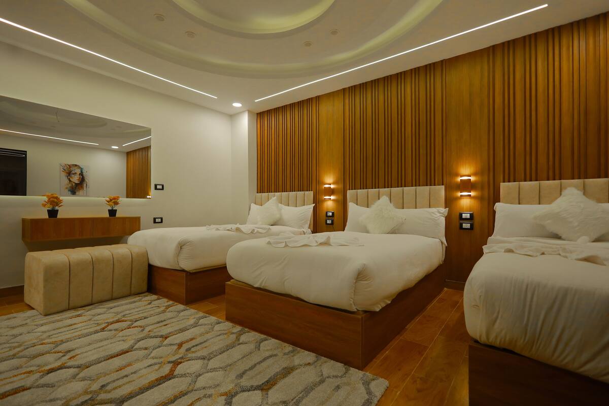 A modern hotel room with three beds, a wooden headboard, a large mirror, and a decorative rug.