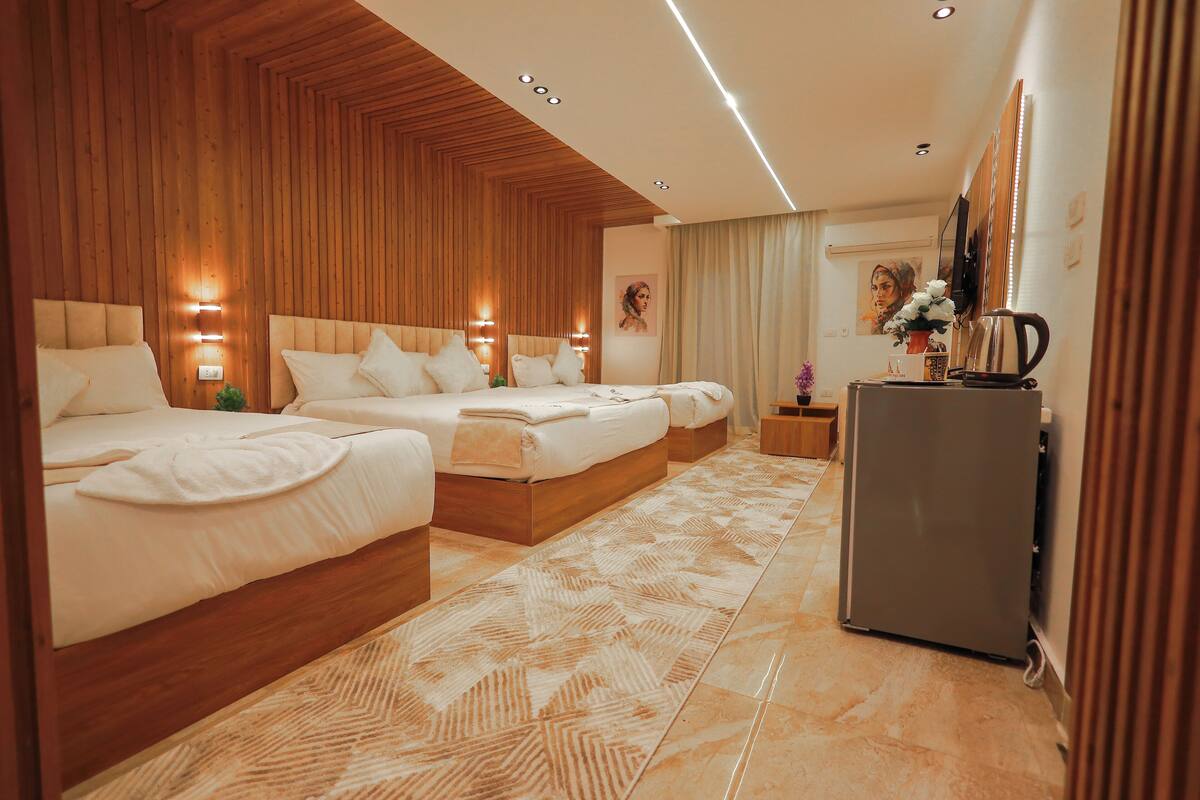 A hotel room with three beds, wooden paneling, a patterned rug, a refrigerator, and a television on the wall.