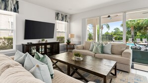 Een moderne woonkamer met een grote flatscreen-tv, een houten salontafel, een beige bank en een groot raam met uitzicht op een zwembad en groen.