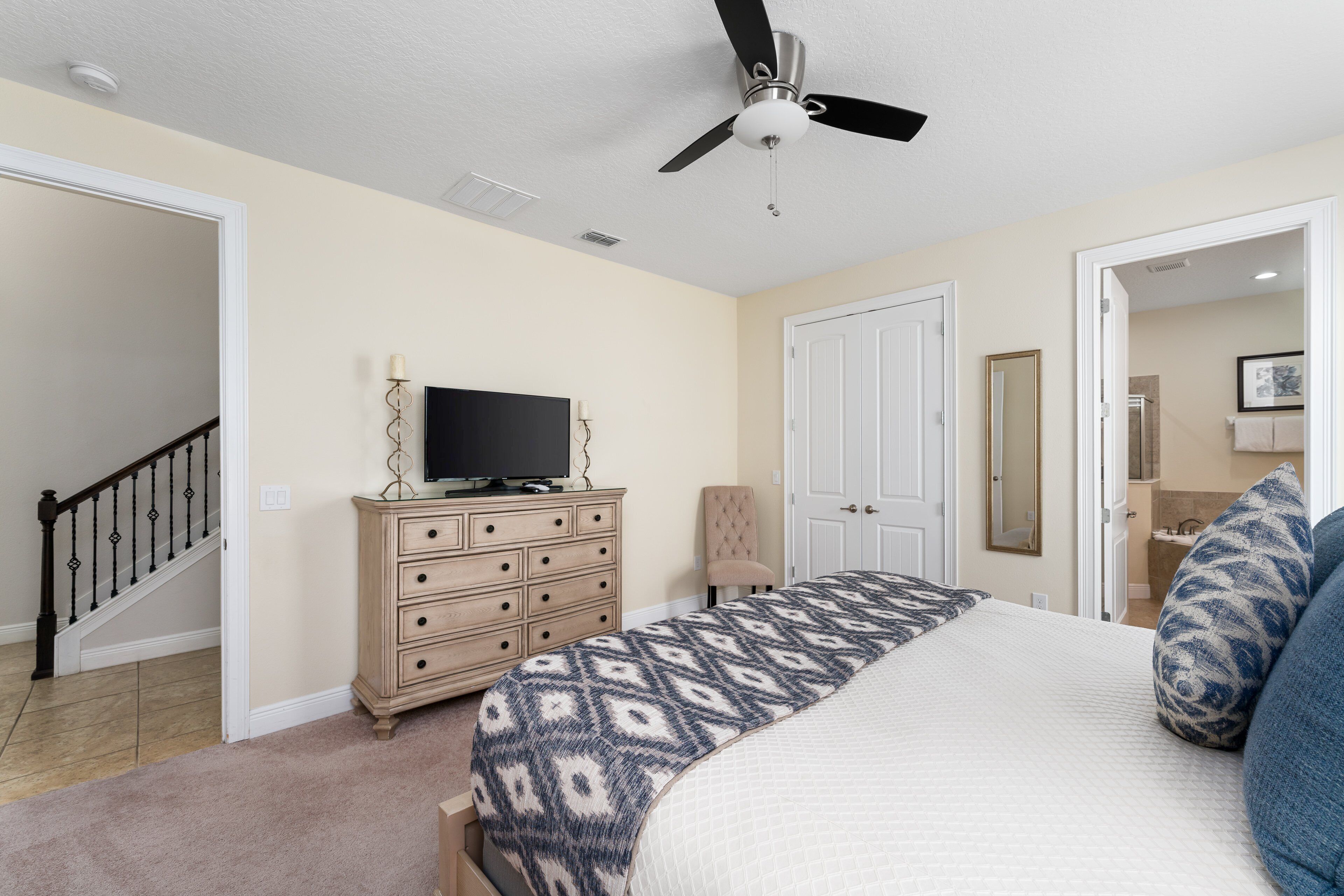Une chambre à coucher comprenant un lit, une commode, un téléviseur, un ventilateur de plafond et une porte donnant sur une salle de bain.