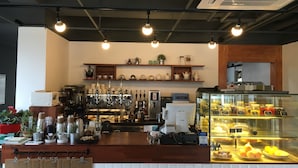 Interior de una cafetería con mostrador, máquinas de café, estantes con vajillas de té y una vitrina con pasteles.