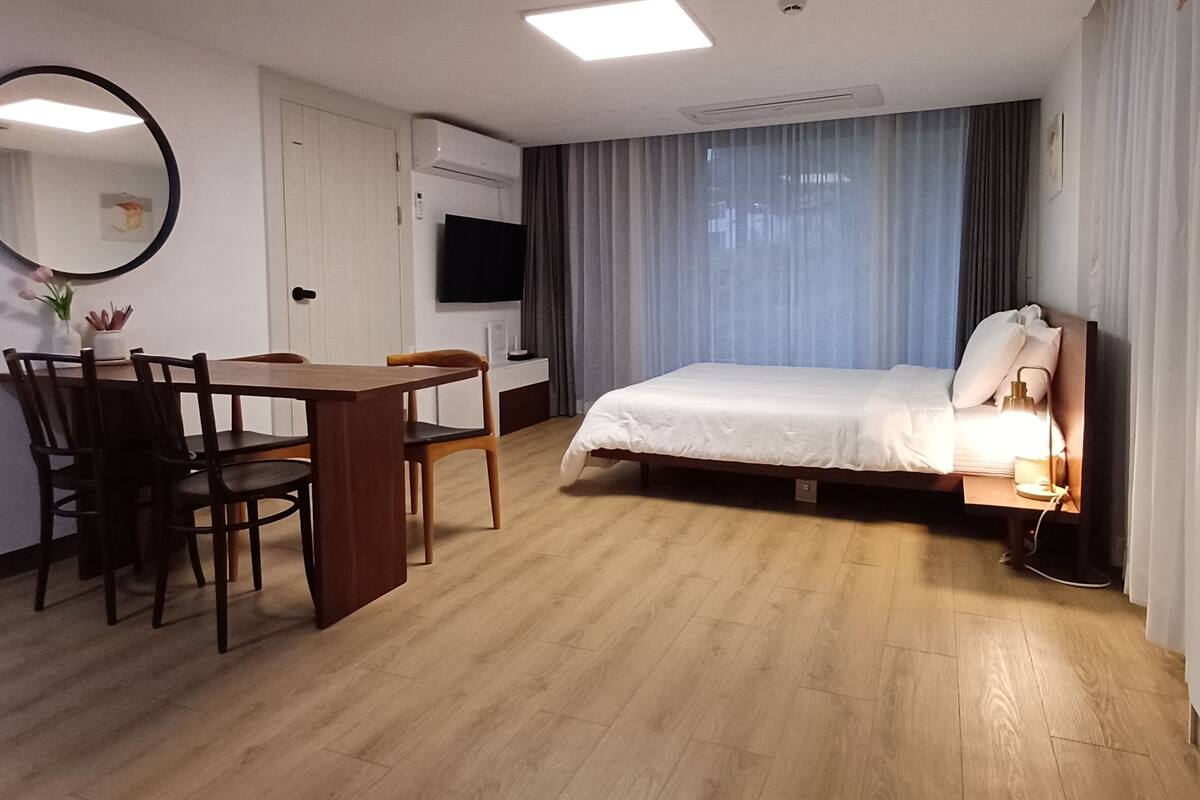A hotel room with a bed, a desk, a chair, a TV, and an air conditioning unit.