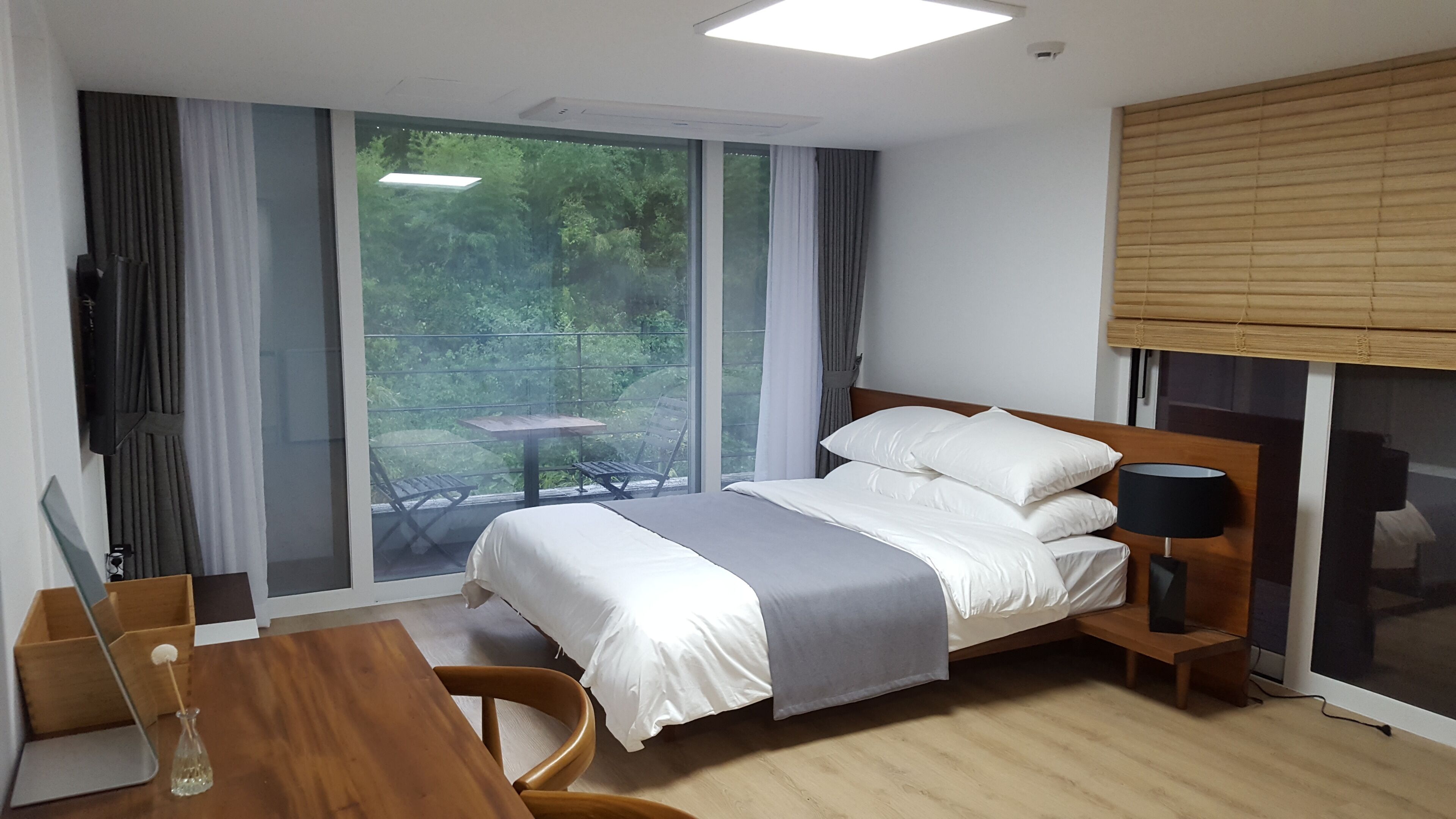 A hotel room with a bed, a desk, a chair, a table, and a window with a view of greenery.
