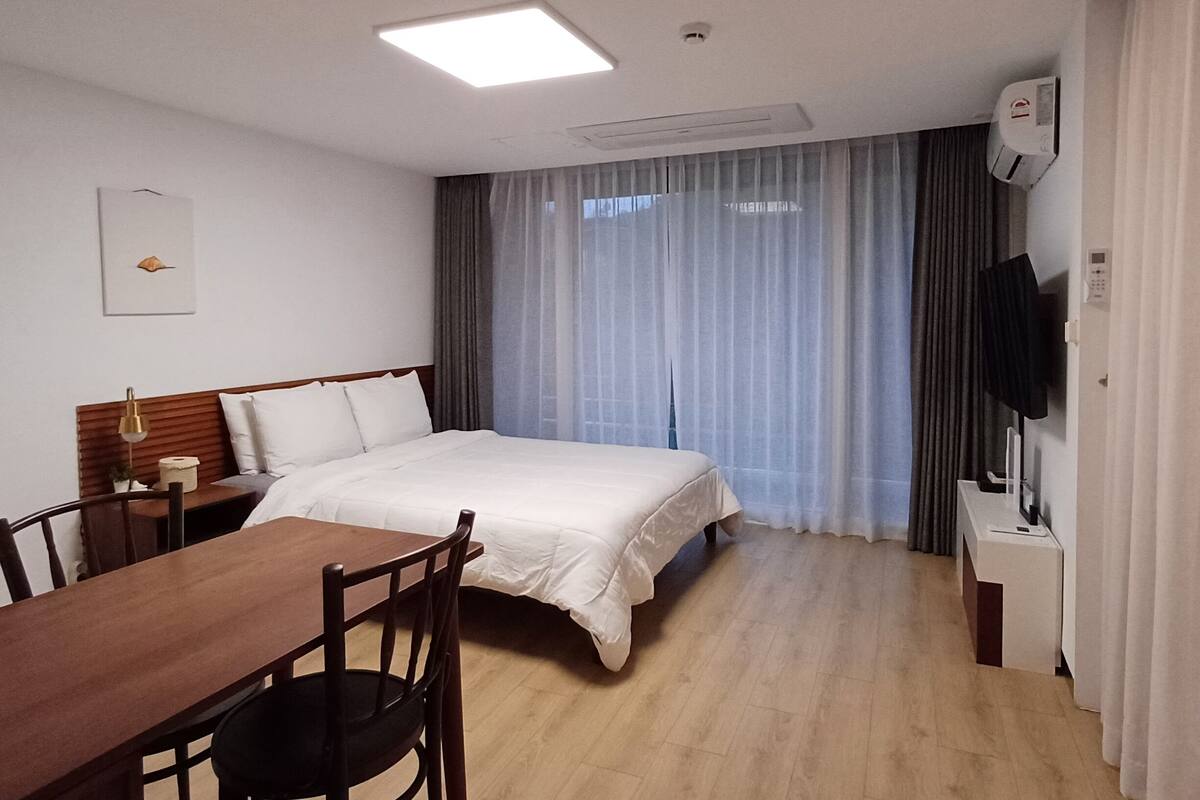 A hotel room with a bed, a desk with a chair, a TV, and a window with curtains.