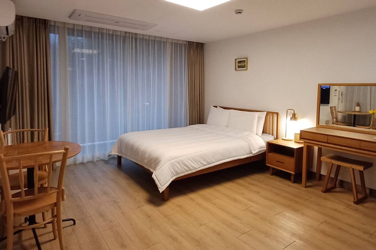 A hotel room with a bed, a desk, a chair, a bedside table, a lamp, a mirror, and a window with curtains.