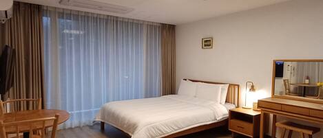 Habitación de hotel con cama, escritorio, silla, mesita de noche, lámpara, espejo y ventana con cortinas.