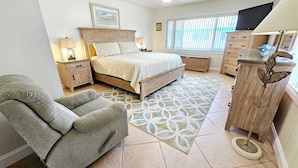 Een slaapkamer met een bed, een stoel, een dressoir, een televisie en een raam met jaloezieën.