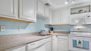 A modern kitchen with white cabinets, a granite countertop, a stainless steel sink, a built-in microwave, and a dishwasher. - Castle Reef 128 Walk to the Ocean (New Smyrna Beach)