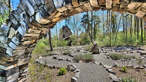 Un arc en pierre encadre un sentier rustique pavé de pierres qui mène à un chalet en bois situé dans une zone boisée.