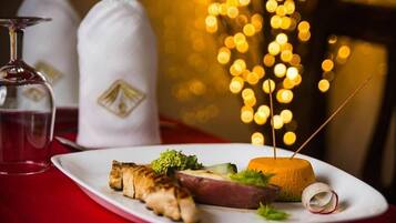 A plate with grilled chicken, a slice of cheese, and a round cheese wheel on a white plate, set on a red table with a glass of water and two white bags in the background.
