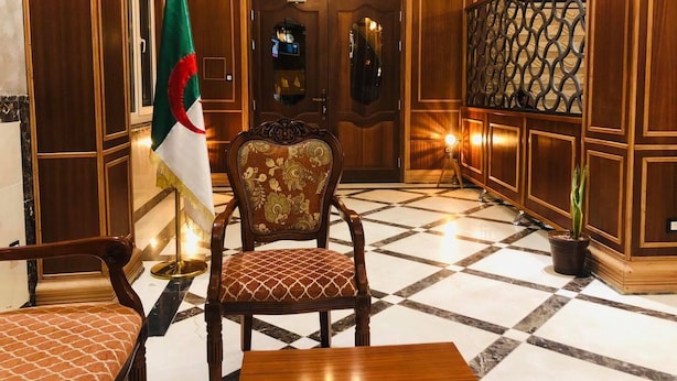 Un vestíbulo con un mostrador de recepción de madera, una bandera, una silla estampada y una escalera con barandilla decorativa.