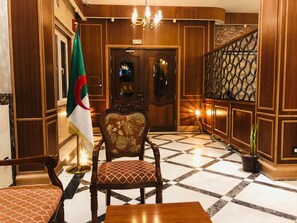 A lobby with a wooden reception desk, a flag, a patterned chair, and a staircase with a decorative railing.