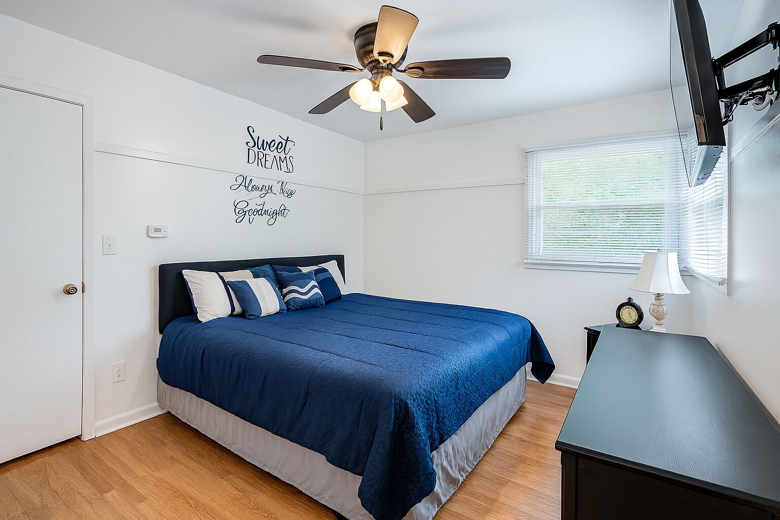 A bedroom with a bed, a ceiling fan, a wall clock, and a window with blinds.