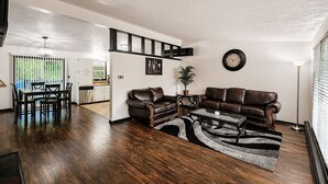 A spacious living room with a dining area, a kitchen in the background, and a large window with blinds. - Serene Family Home: Sleeps 16! (Wyoming)